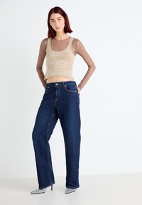 Débardeur beige avec un motif discret, superposé sous une chemise à manches longues en voile marron. Jean en denim bleu foncé à jambe large, talons métalliques pointus.