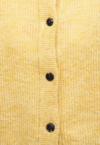 Žuta pletena tkanina s vertikalnim rebrastim detaljem, koja sadrži tri tamna okrugla dugmeta ravnomjerno raspoređena duž središnjeg šava.