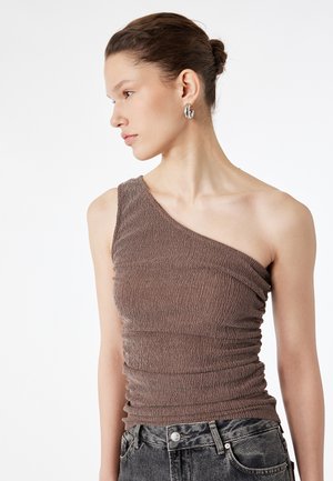 Jeune femme avec les cheveux attachés en chignon, portant des boucles d'oreilles argentées en forme d'anneau, un haut texturé marron à une épaule, et un jean noir délavé taille haute, regardant vers la gauche.