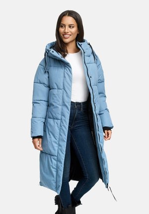 Vrouw in een lange lichtblauwe gewatteerde jas met capuchon, witte top, donkerblauwe jeans en zwarte laarzen, staand en lachend tegen een witte achtergrond.