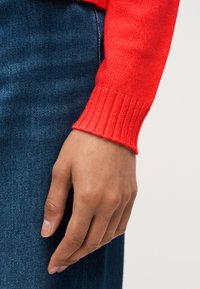 Χέρι ακουμπισμένο στο πλάι, φορώντας φωτεινό κόκκινο πλεκτό μανίκι πουλόβερ και μπλε τζιν, πάνω σε ουδέτερο φόντο.
