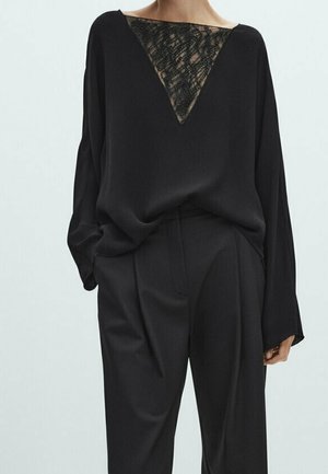 Personne portant un haut noir ample à manches longues avec une incrustation triangulaire en dentelle au niveau de l'encolure et un pantalon noir taille haute assorti.
