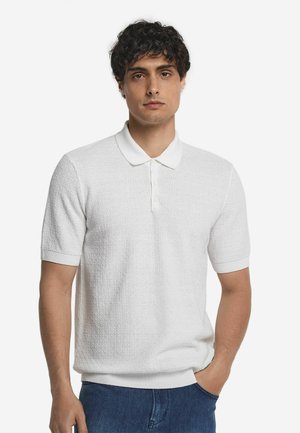 Vit kortärmad polo med ett texturerat stickmönster, med klassisk krage och ribbade manschetter. Matchas med blå jeans.
