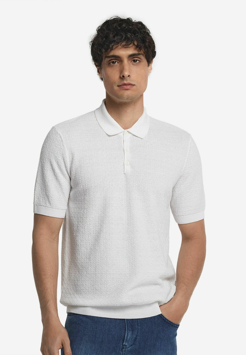 Vit kortärmad polo med ett texturerat stickmönster, med klassisk krage och ribbade manschetter. Matchas med blå jeans.