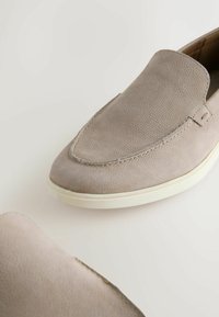 Chaussure à enfiler en daim beige clair avec semelle blanche, bout arrondi et coutures détaillées sur une tige en cuir lisse.