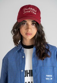 Căciulă de baseball roșie cu „Club Social Interzis” brodată în alb. Asortată cu o cămașă cu carouri albastre și un tricou grafic alb.