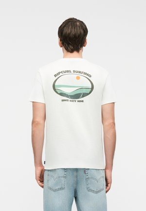Man draagt een wit t-shirt met een Ripcurl Surfing-logo en golfgrafiek, gecombineerd met lichtblauwe jeans, gefotografeerd van achteren.