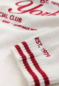 Tissu tricot blanc avec texte brodé rouge "EST. 1979," rayures rouges sur le bord-côte et lettres rouges partiellement visibles.