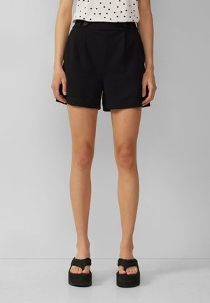 Vrouw draagt zwarte shorts met hoge taille en knopen en zwarte platformsandalen, gecombineerd met een wit mouwloos topje met zwarte stippen.