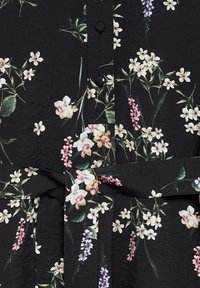 Blouse en tissu noir avec divers motifs floraux en blanc, rose et violet. Elle possède une patte de boutonnage et un détail de taille à nouer.