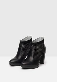 Para czarnych skórzanych botków za kostkę na blokowych obcasach z perforowanymi siateczkowymi bokami, wyeksponowanych na białej powierzchni na tle jasnoszarym.