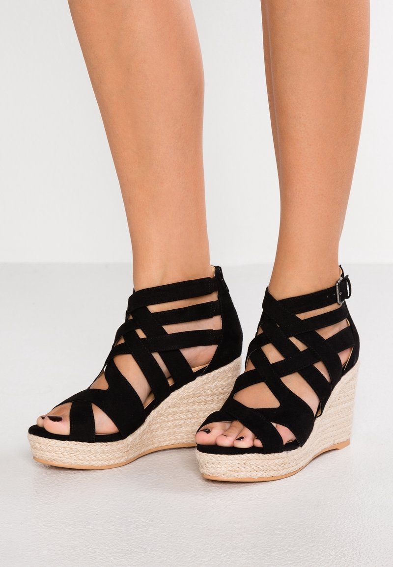 Jambes portant des sandales compensées noires à brides avec des talons espadrilles tissés et des boucles à la cheville sur un sol gris clair.