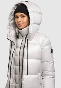 Ezüst színű tollkabát magas gallérral és nagy kapucnival, cipzáras elülső résszel és fekete húzózsinórokkal. Fekete logó a ujján.