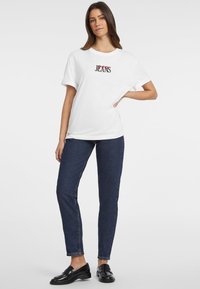 T-shirt bianco in cotone con grafica "JEANS" in rosso e nero, abbinata a jeans a vita alta blu scuro e mocassini neri. Design semplice e casual.