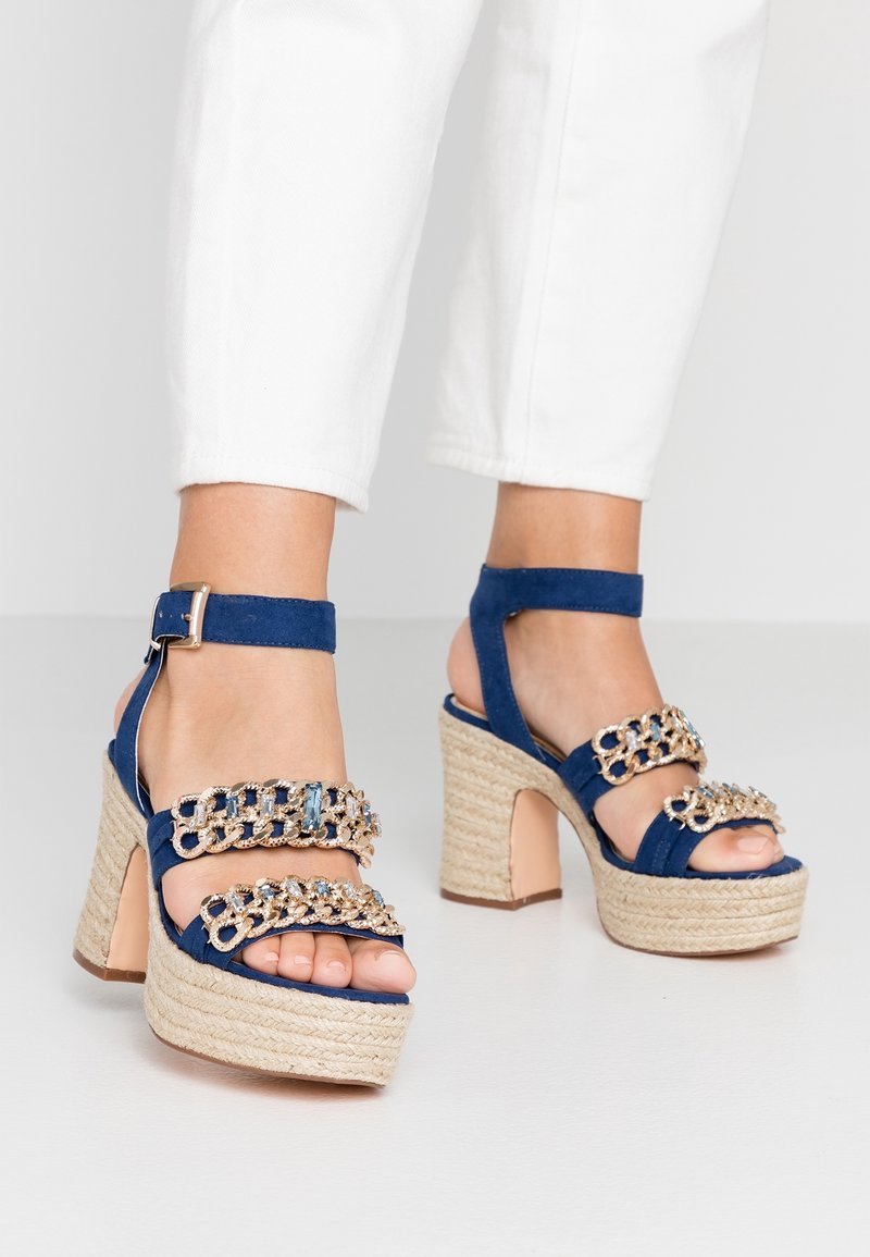 Pieds portant des sandales à talons hauts compensés bleues avec semelles en jute tressée et brides décoratives en chaîne dorée, assorties à un pantalon blanc court.