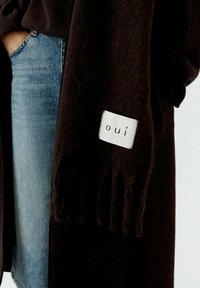 Schwarzer Wollschal mit Fransen und einem weißen Etikett mit der Aufschrift "oui", kombiniert mit hellblauer Jeans. Strukturiertes Material, lässige Designdetails.