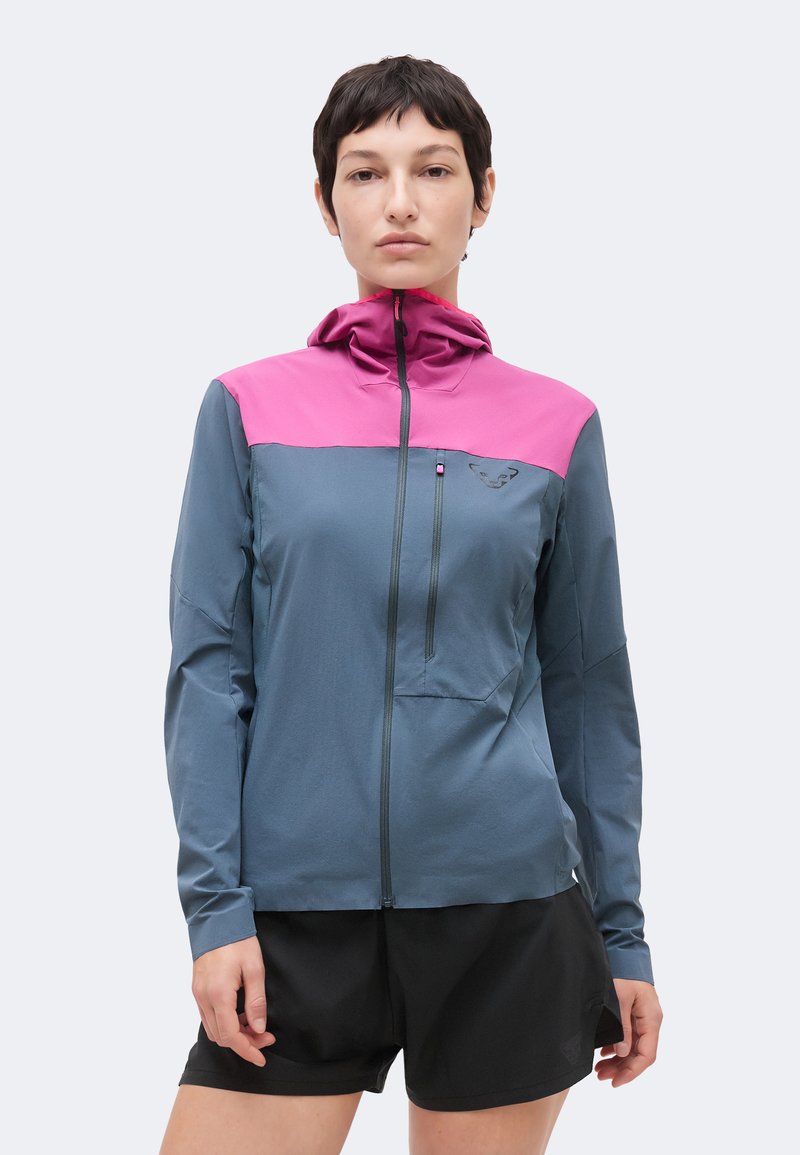 Femme portant une veste à capuche zippée bleue et rose avec une petite poche poitrine, associée à un short noir, debout devant un fond uni.