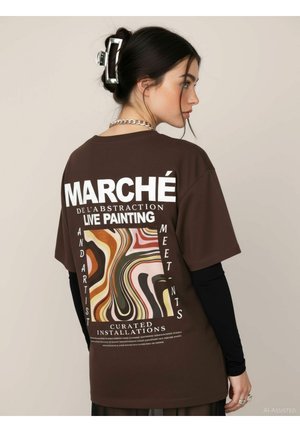 Ung kvinde med mørkt hår i en clips iført en brun T-shirt med hvid tekst og et abstrakt flerfarvet bølget mønster på ryggen.