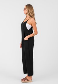 Jumpsuit nero senza maniche con una vestibilità rilassata, caratterizzato da tasche laterali e un design delle gambe leggermente affusolato. Il modello indossa sandali color sabbia e occhiali da sole.