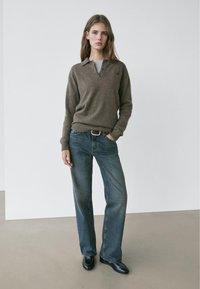 Bruine, gebreide trui met een kraag, gecombineerd met losse, blauwe spijkerbroek en zwarte schoenen, staand tegen een neutrale achtergrond.