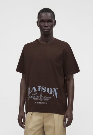Bărbat cu păr scurt purtând un tricou maro supradimensionat cu text mare gri "Maison Margiela" și pantaloni bej pe un fundal simplu.