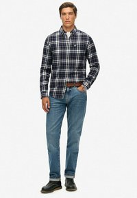 Marineblauw-wit geruit overhemd met lange mouwen, gecombineerd met een blauwe spijkerbroek en zwarte schoenen. Opvallende details zijn de knoopsluiting bij de kraag en het getailleerde ontwerp.