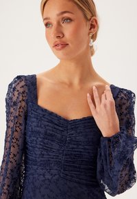 Marindress med texturerad, draperad bodice och genomskinliga, broderade puffärmar; har en fyrkantig halsringning och delikata detaljer.