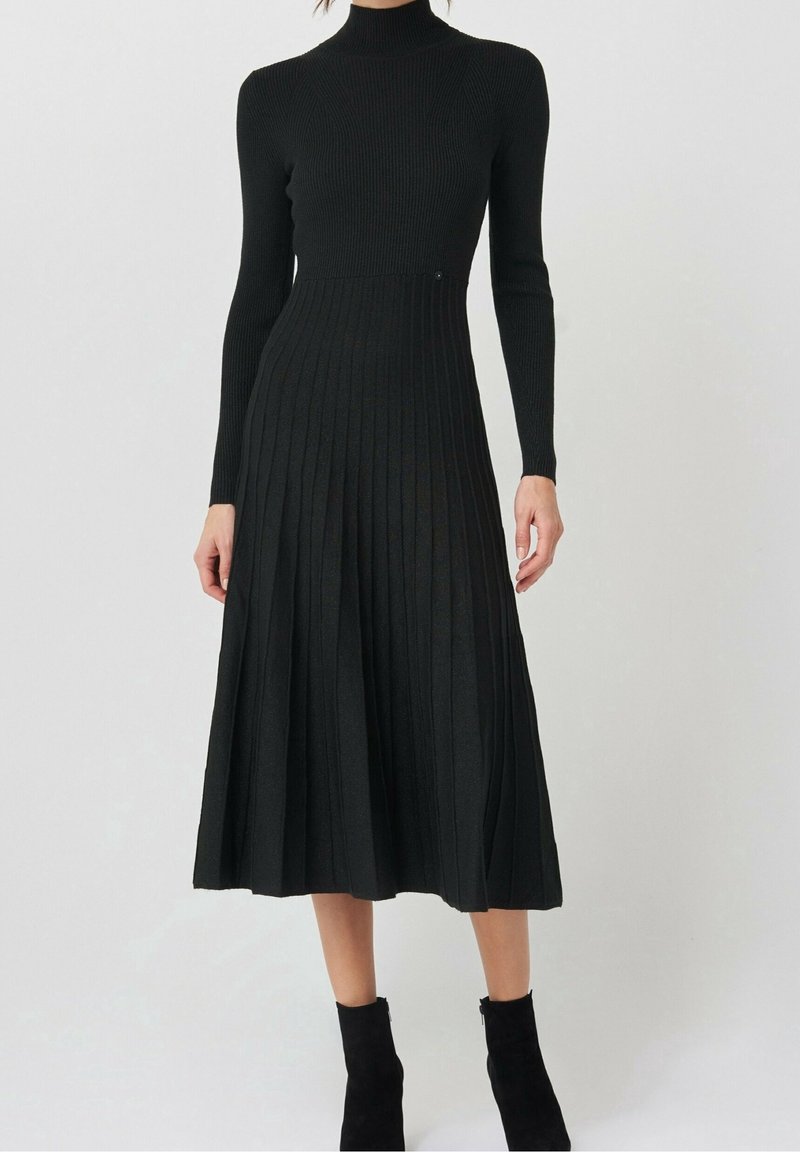 Femme portant une robe midi noire à manches longues en maille côtelée avec une jupe plissée et des bottines noires, debout devant un fond uni.