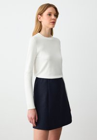 Weißes langärmeliges Oberteil, kombiniert mit einem marineblauen Faltenrock mit vertikalen Streifen, glatter Textur und taillierter Silhouette.