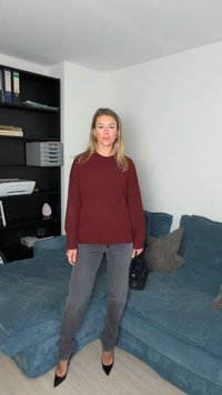 Bordo džemper s dugim rukavima, sive traperice i crne cipele na visoku petu, stojeći na plavom sofama od kordurovog materijala s jastučićima s uzorkom.