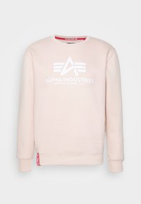 Felpa rosa chiaro con girocollo, logo e testo bianchi di Alpha Industries, polsini e orlo a coste, e etichetta rossa vicino al lato sinistro in basso.