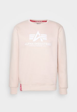 Felpa rosa chiaro con girocollo, logo e testo bianchi di Alpha Industries, polsini e orlo a coste, e etichetta rossa vicino al lato sinistro in basso.