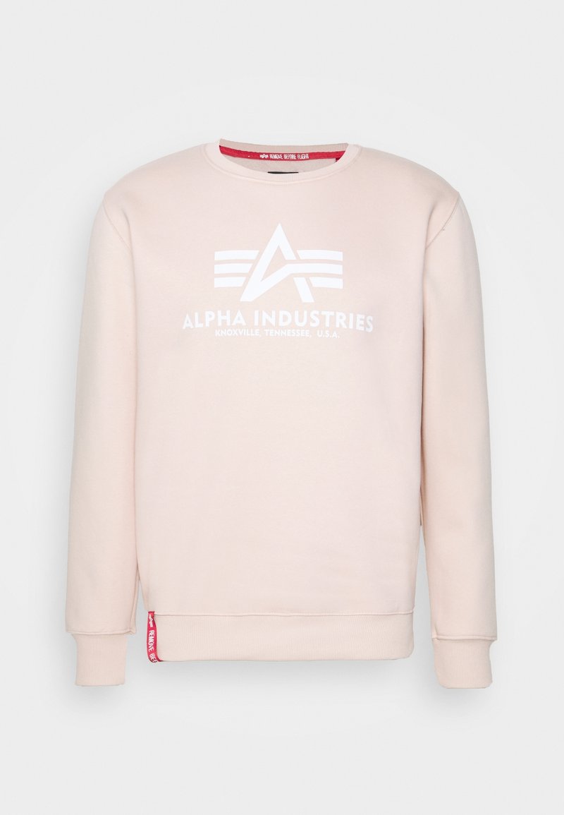 Felpa rosa chiaro con girocollo, logo e testo bianchi di Alpha Industries, polsini e orlo a coste, e etichetta rossa vicino al lato sinistro in basso.