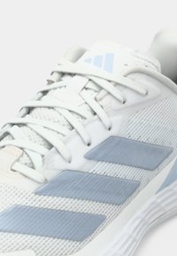 Chaussure de sport blanche avec tige en mesh, accents bleu clair et trois rayures grises. Design à lacets avec détails texturés sur la semelle.