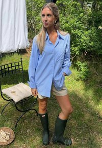 Femme portant une chemise rayée bleue, un short beige et des bottes vert foncé, debout à l'extérieur près d'une chaise noire avec coussin et du linge qui sèche.