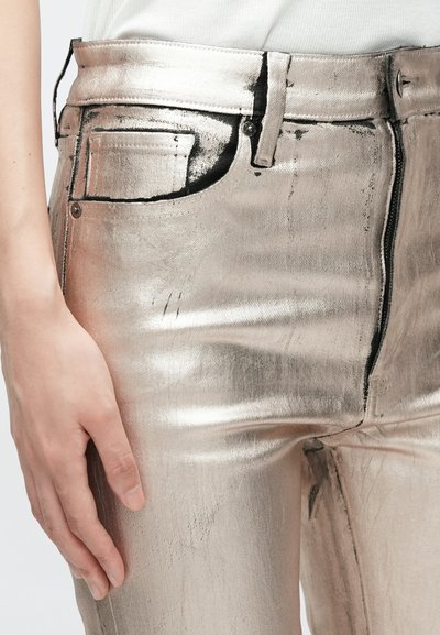 Jeans skinny argent métallique à la texture lisse, avec poches avant, couture visible et fermeture éclair. Main reposant sur le côté gauche.