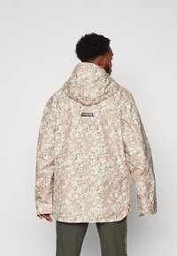 Veste camouflage beige avec des motifs floraux blancs et verts, dotée d'une capuche, de manches longues et d'une étiquette avec logo au dos.