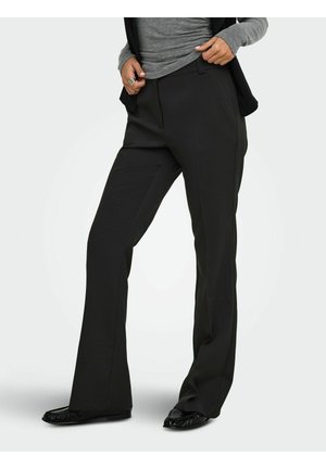 Personne portant un pantalon noir droit avec des poches avant, un haut gris à manches longues et des chaussures plates noires, debout devant un fond uni.