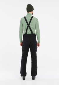 Zwarte skibroek met verstelbare bretels, gladde textuur en met rits sluitende zakken. Gepaarde met een groene lange mouwen shirt en een beanie.