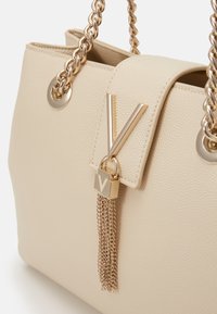 Beige Handtasche aus Kunstleder mit Kettengriffen, goldfarbenen Beschlägen, V-förmigem Akzent und Quasten-Detail, das eine strukturierte Oberfläche aufweist.