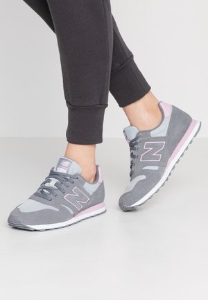 Personne portant des baskets New Balance grises et roses avec un pantalon de jogging noir, debout avec un pied légèrement levé.