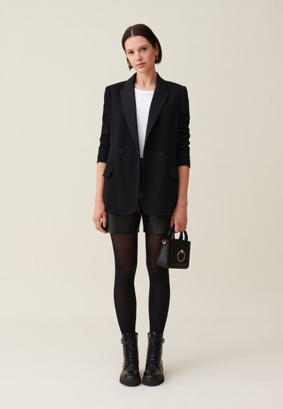 Blazer nero su una maglietta bianca, abbinato a pantaloni in pelle nera, collant opachi e stivali combat neri. Mini borsa nera con dettaglio.