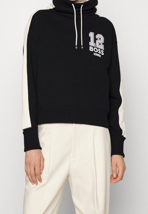 Schwarz-weißes Kapuzenpullover mit der Aufschrift "12 BOSS ICONIC" auf der Brust, weißen Kordeln und weißen Seitenstreifen, getragen zu weißen Hosen.