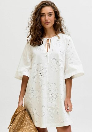 Göndör hajú nő, aki fehér, virágos lyukacsos ruhát visel, bal kezében mintás barna dzsekit tart.