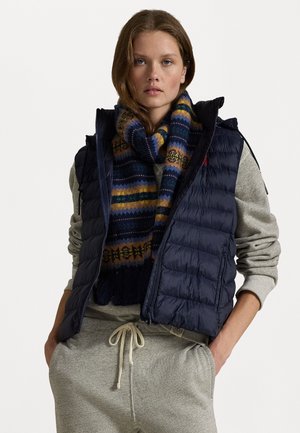 Gilet imbottito blu navy con chiusura a zip, dotato di cappuccio e un sottile design trapuntato. Abbinato a una sciarpa colorata con motivo e a una felpa grigia.
