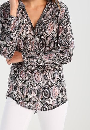 Frau trägt ein langärmliges Hemd mit geometrischem Sechseckmuster in Schwarz-, Weiß- und Rosatönen, kombiniert mit weißen Hosen.