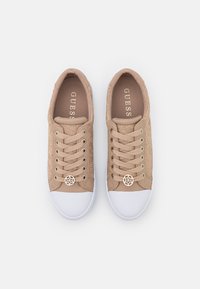 Paire de baskets beiges en dentelle avec bouts en caoutchouc blanc, lacets beiges et breloques métalliques avec logo de la marque sur fond blanc.