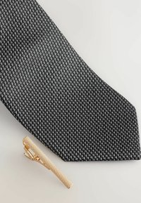 Stribet sort/hvid slips i mønster med tekstureret stof, parret med en guldbelagt slipsenål med ribbet design. Begge dele ligger på en lys overflade.