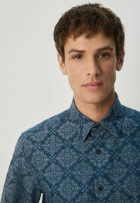 Modrá košeľa s vzorom, ktorá má dekoračný potlač, golier s gombíkmi a tmavé gombíky, vyrobená z hladkej látky s textúrovaným vzhľadom.