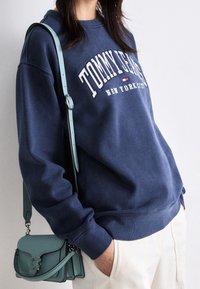 Laivansinis puuvillapaita, jossa on valkoinen "Tommy Jeans" -logo. Yhdistetty vaaleansiniseen olkalaukkuun, joka on varustettu soljella ja säädettävällä hihnalla.